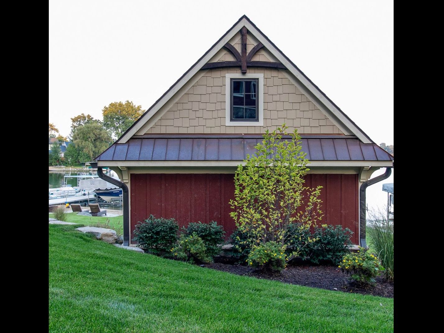 A small red and tan boathouse with a peaked roof, small window, and landscaping.