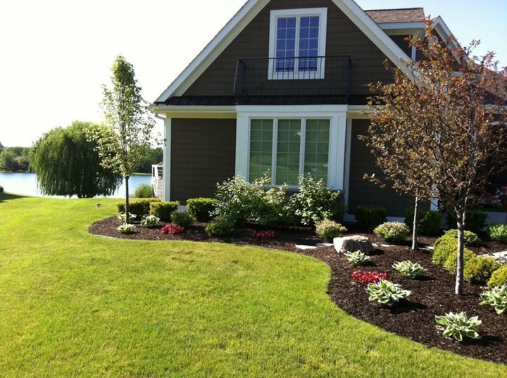 House with landscaped yard, lake in background. Brown house, green lawn, flower beds with mulch.