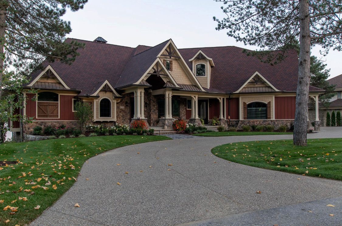 Brown-roofed house with stone and red siding, curved gravel driveway, surrounded by green lawn and trees.