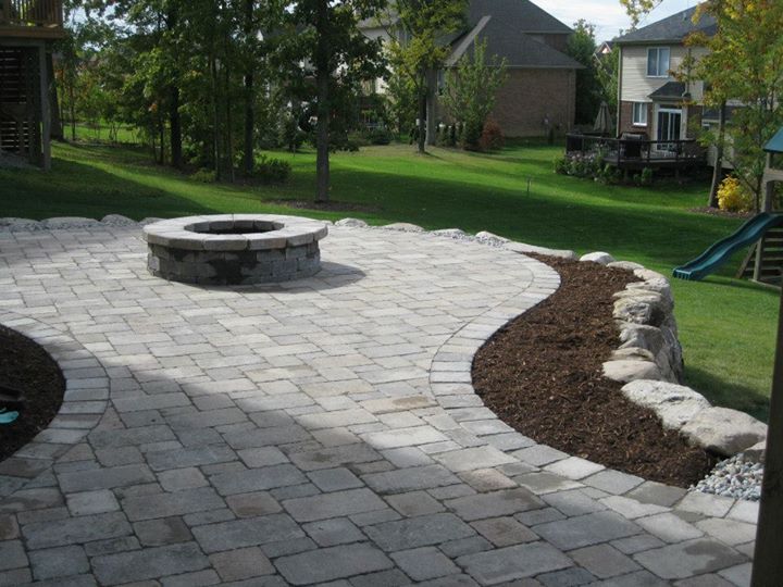 Brick patio with fire pit, bordering garden bed, sloped lawn, and houses in the background.