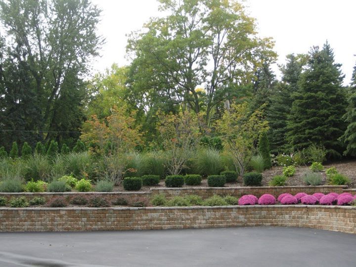 Brick retaining wall with tiered landscaping, featuring shrubs, grasses, and pink flowers against a backdrop of trees.