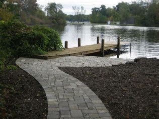 Stone path curves toward a wooden dock on a calm body of water with trees in the background.