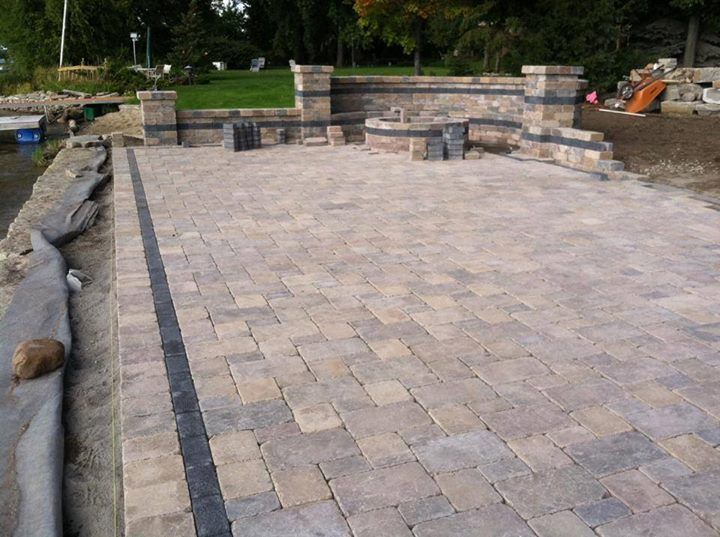 Stone patio with dark border being built near water, with stone wall in the background.