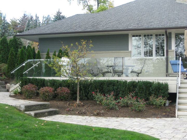 A house with a deck, glass railing, and landscaping. Stone pathway leads to steps. Green grass and trees are in the yard.