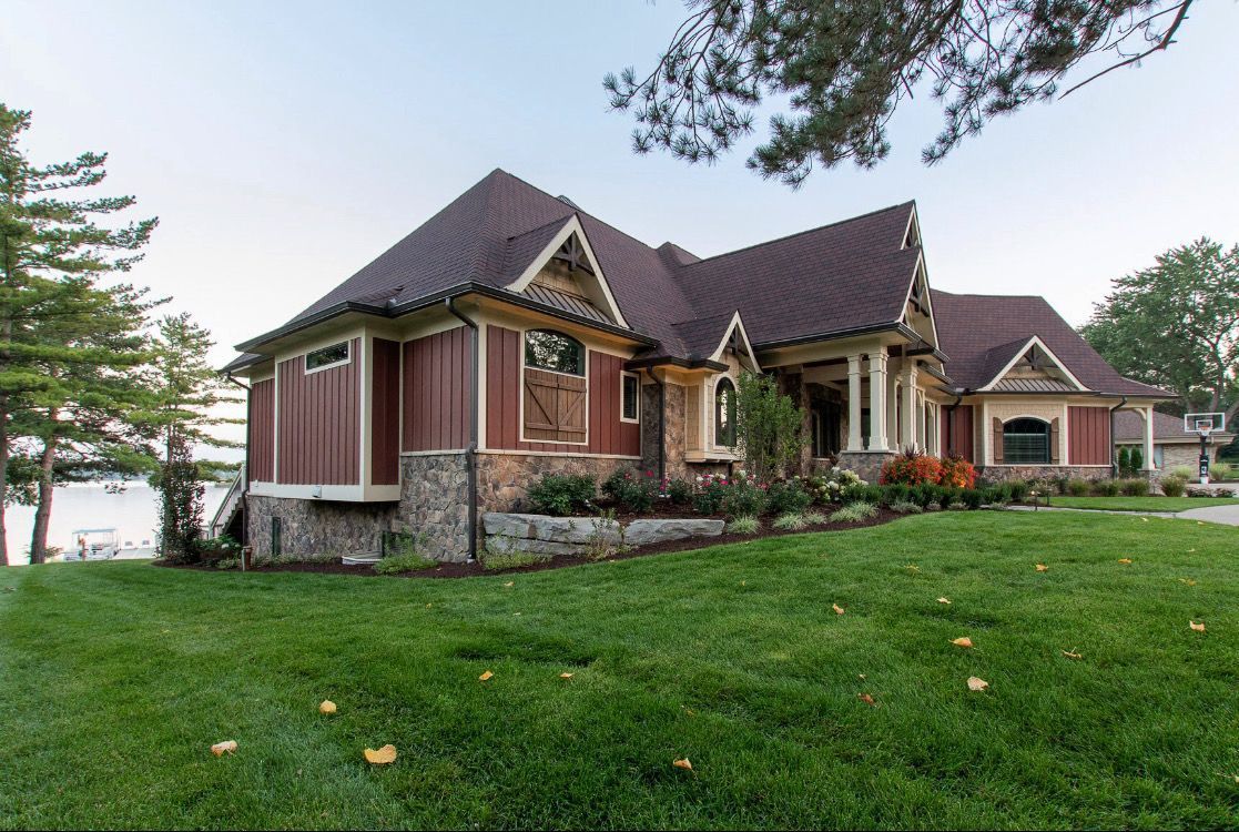 Cottage-style home with red siding, stone accents, and brown roof, situated on a green lawn near water.