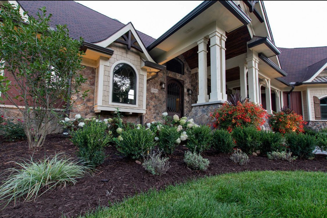 Stone house with a landscaped yard; white columns, arched window, and colorful flowers.