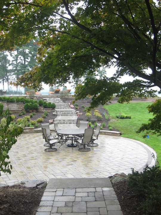 Paved patio with round table and chairs, leading to steps and terraced landscaping. Green lawn and trees surround.