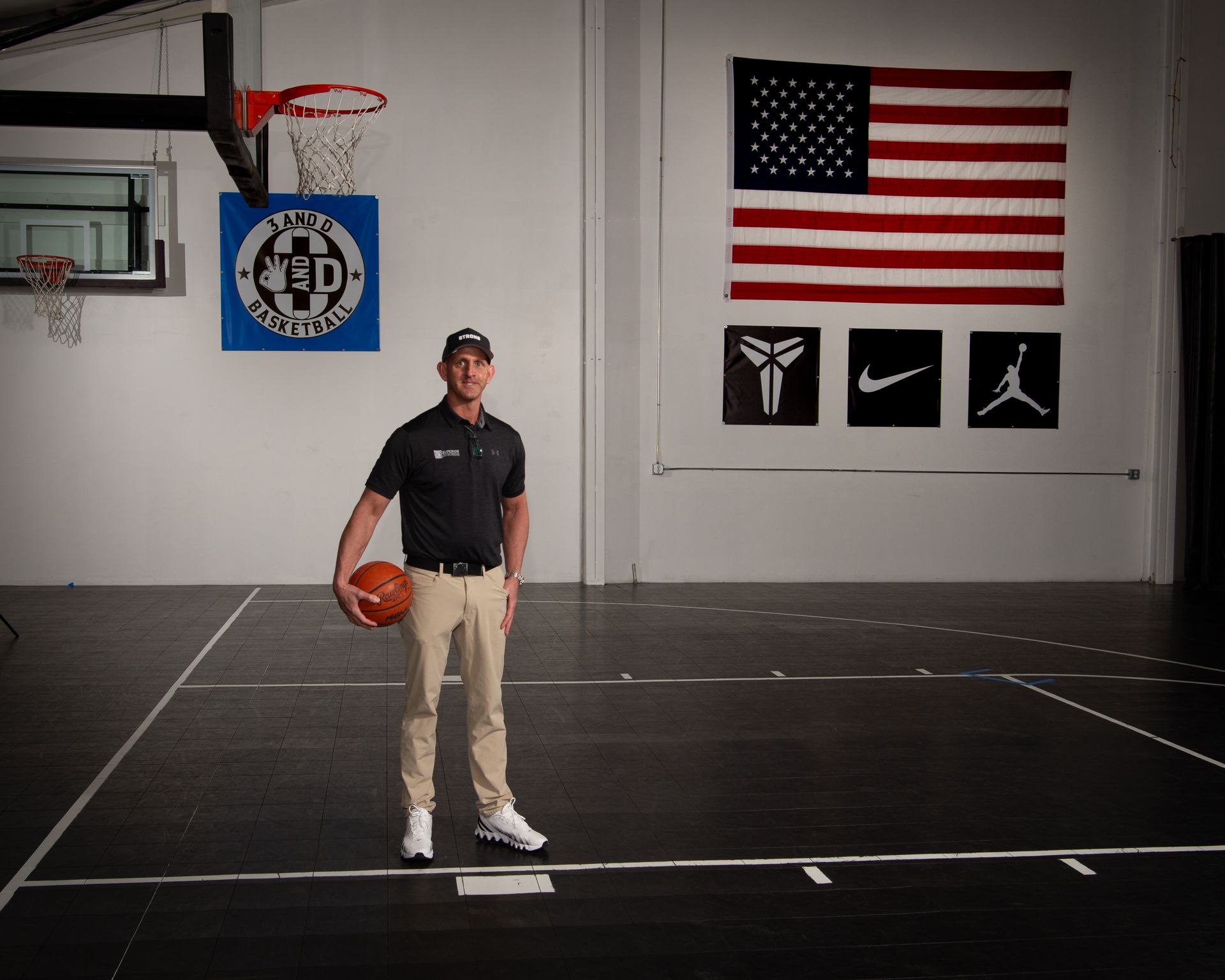 Man holding a basketball on a court, wearing a cap and khaki pants. American flag and logos in the background.