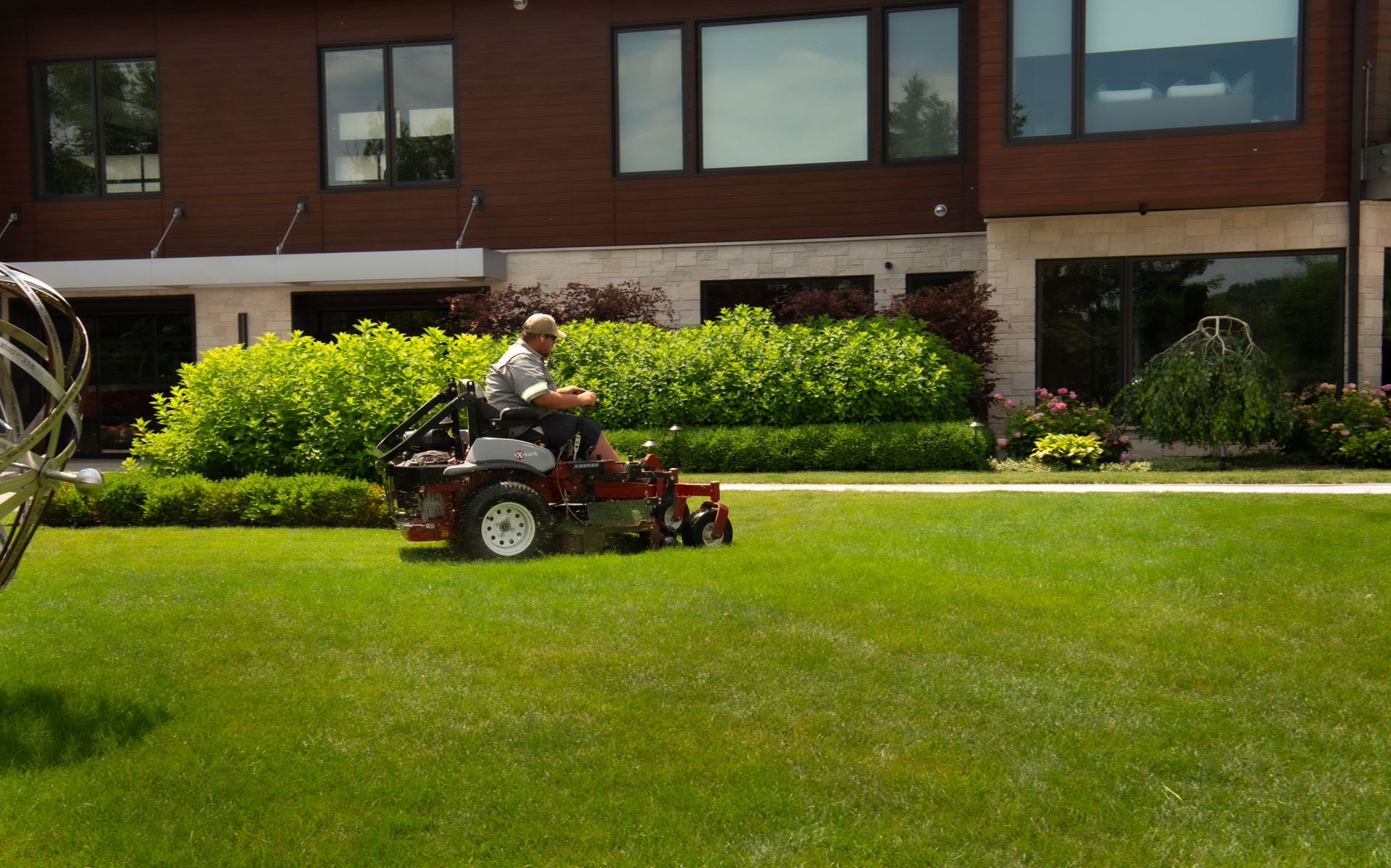 Man on riding lawnmower cutting grass in front of a large house with green bushes and windows.