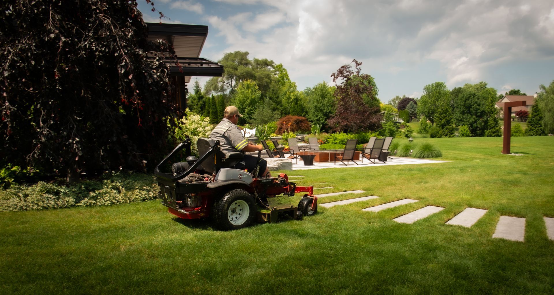 Man mowing a green lawn with a riding mower, stone walkway and outdoor seating area in the background.