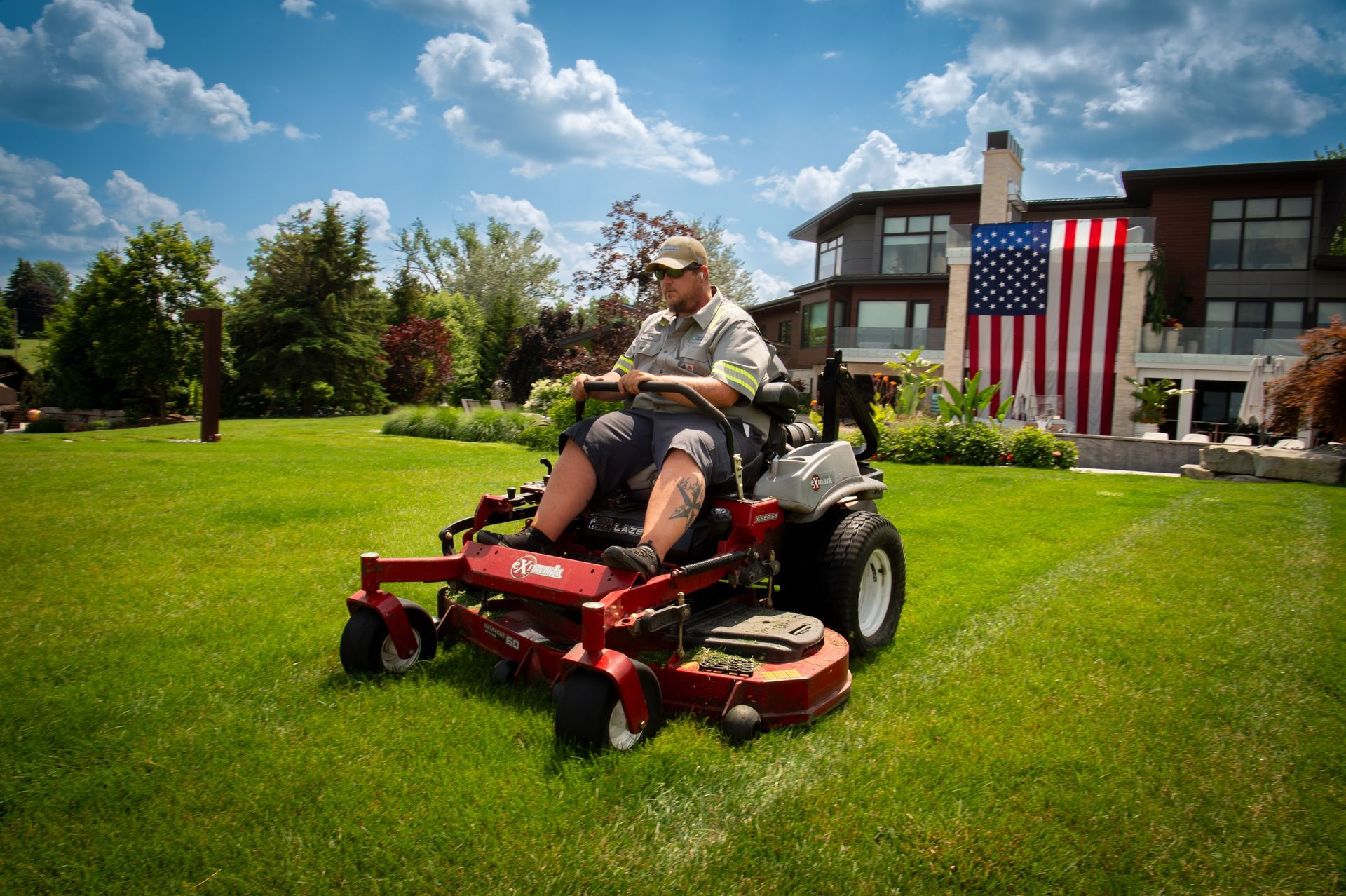 Person on a red zero-turn mower cutting grass in front of a house with a large American flag.