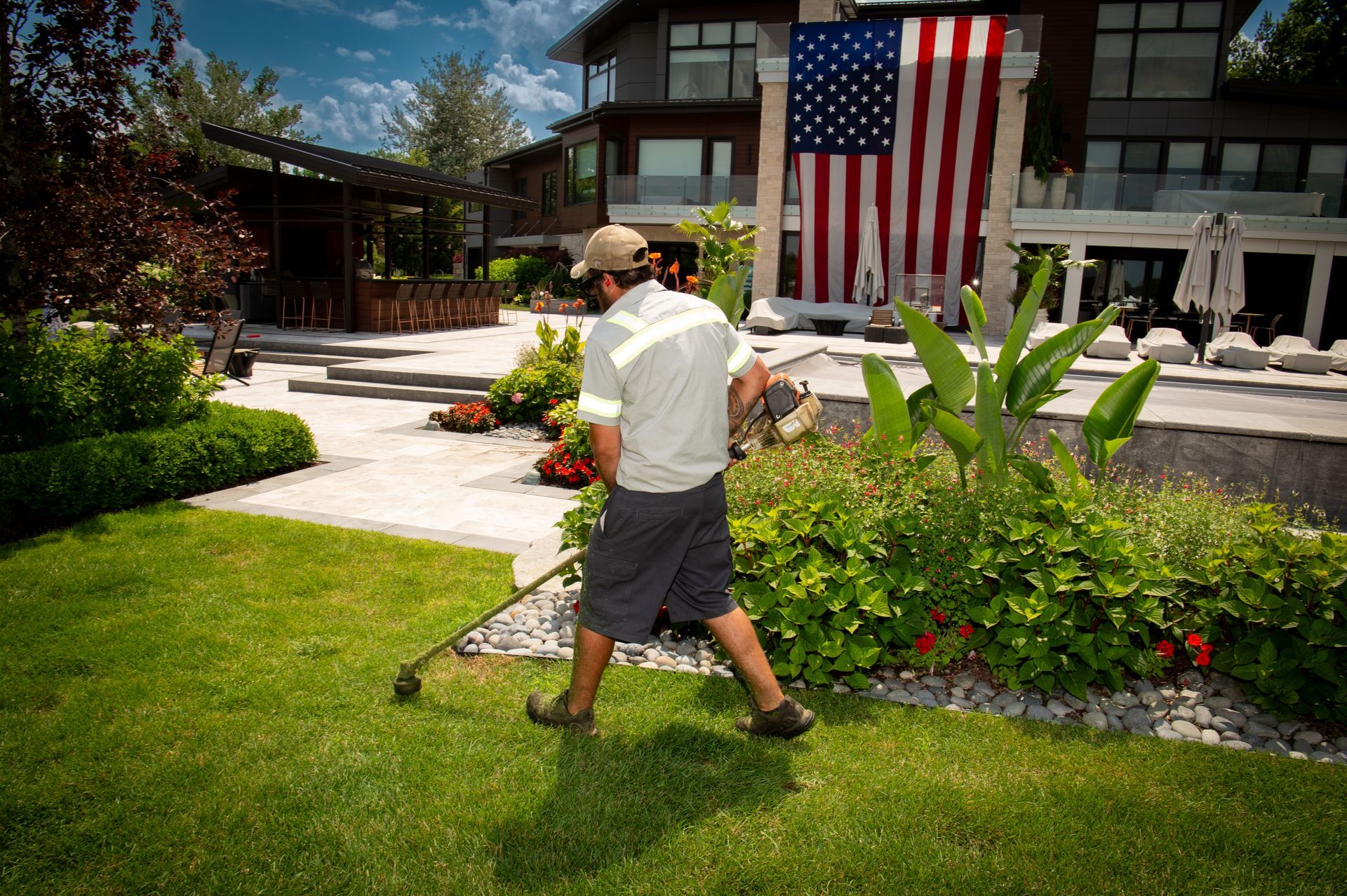 Man trimming grass in a yard with a large house and American flag in the background.