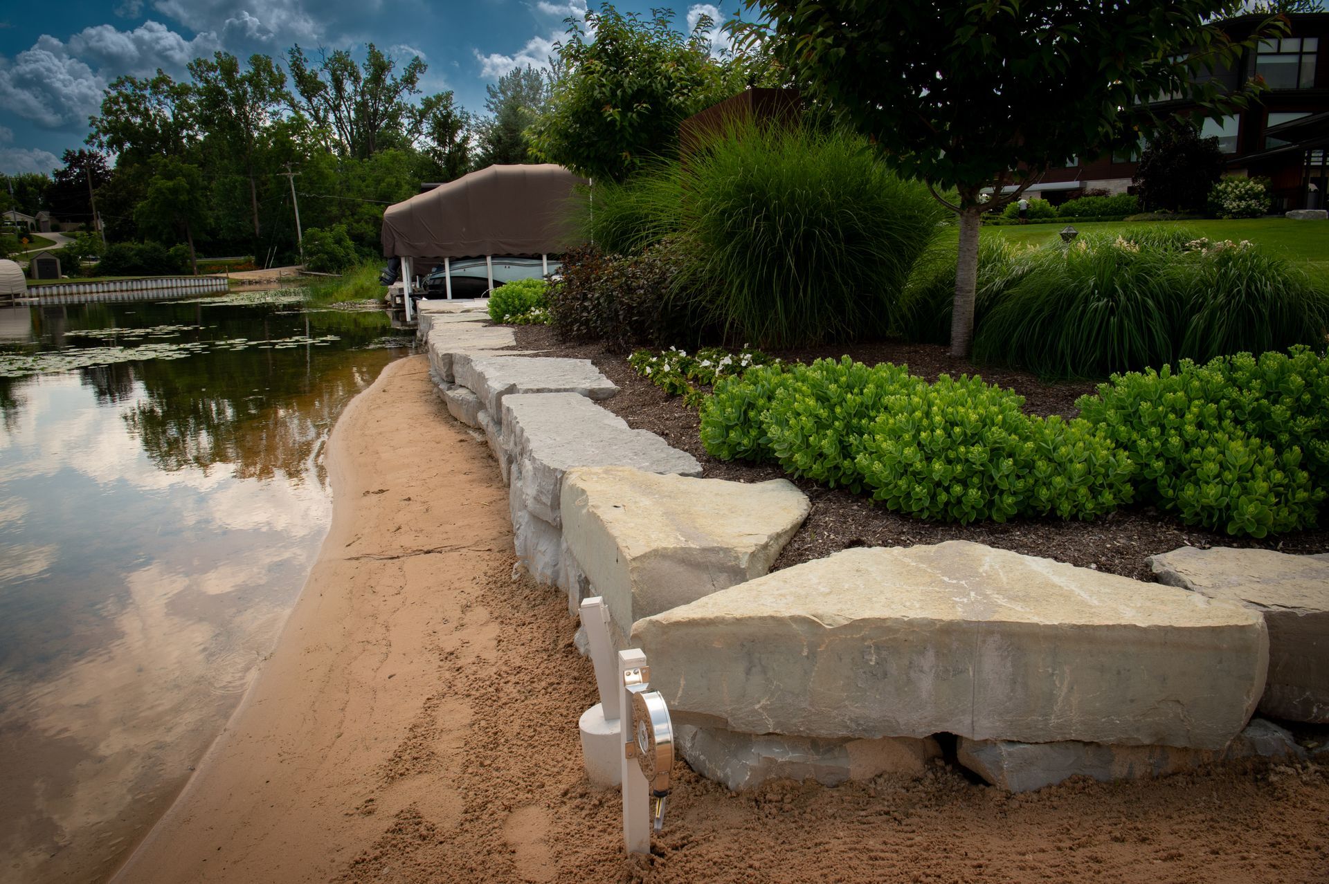 Sandy beach with a stone retaining wall, lush green plants, and a boat lift on a lake.