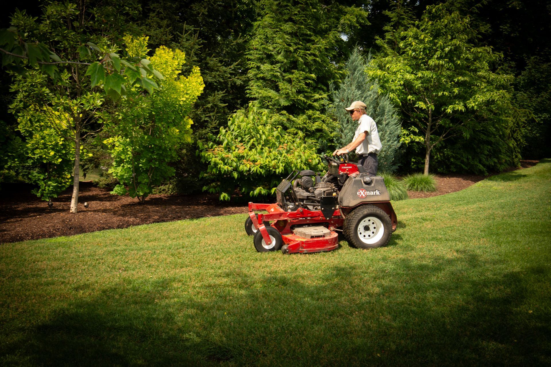 Man mowing a lawn with a red zero-turn mower in a yard lined with trees and mulch.
