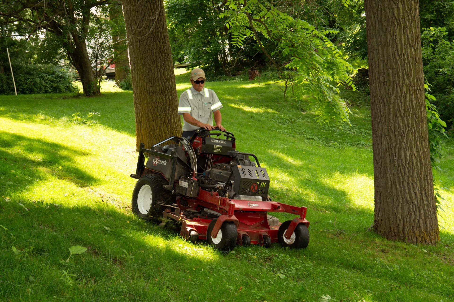 A person operating a red riding lawnmower on a grassy hill, next to trees.