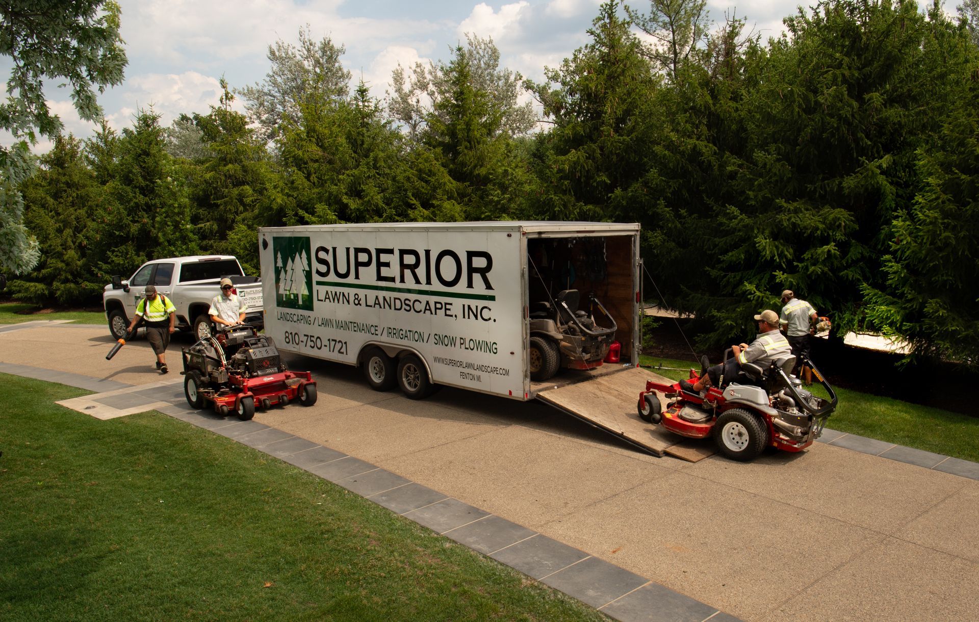 Workers unloading lawnmowers from a trailer on a driveway; a white truck is nearby.
