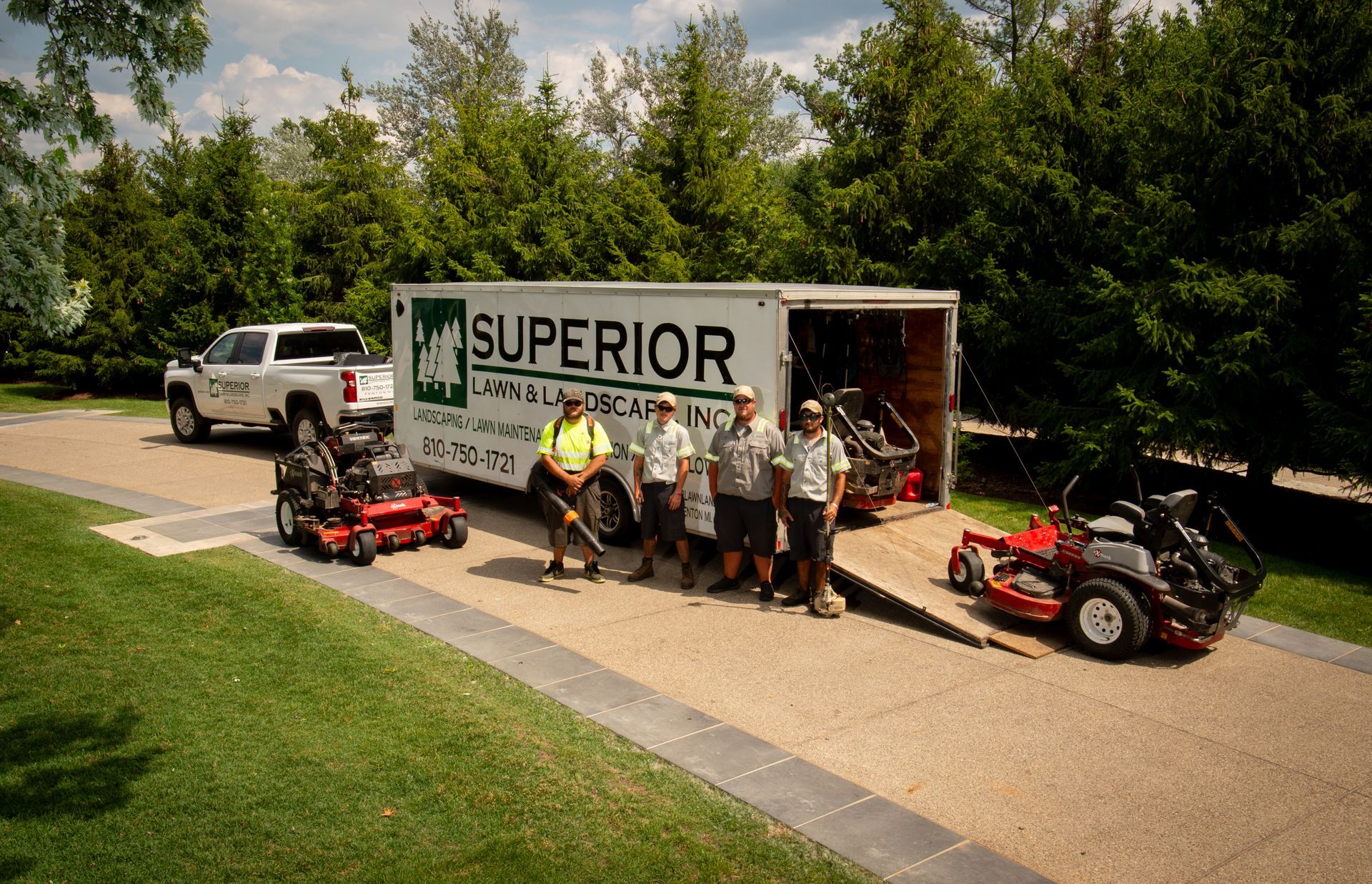 Lawn care crew with truck and equipment. Crew stands in front of a trailer with lawnmowers. Sunny outdoor setting.
