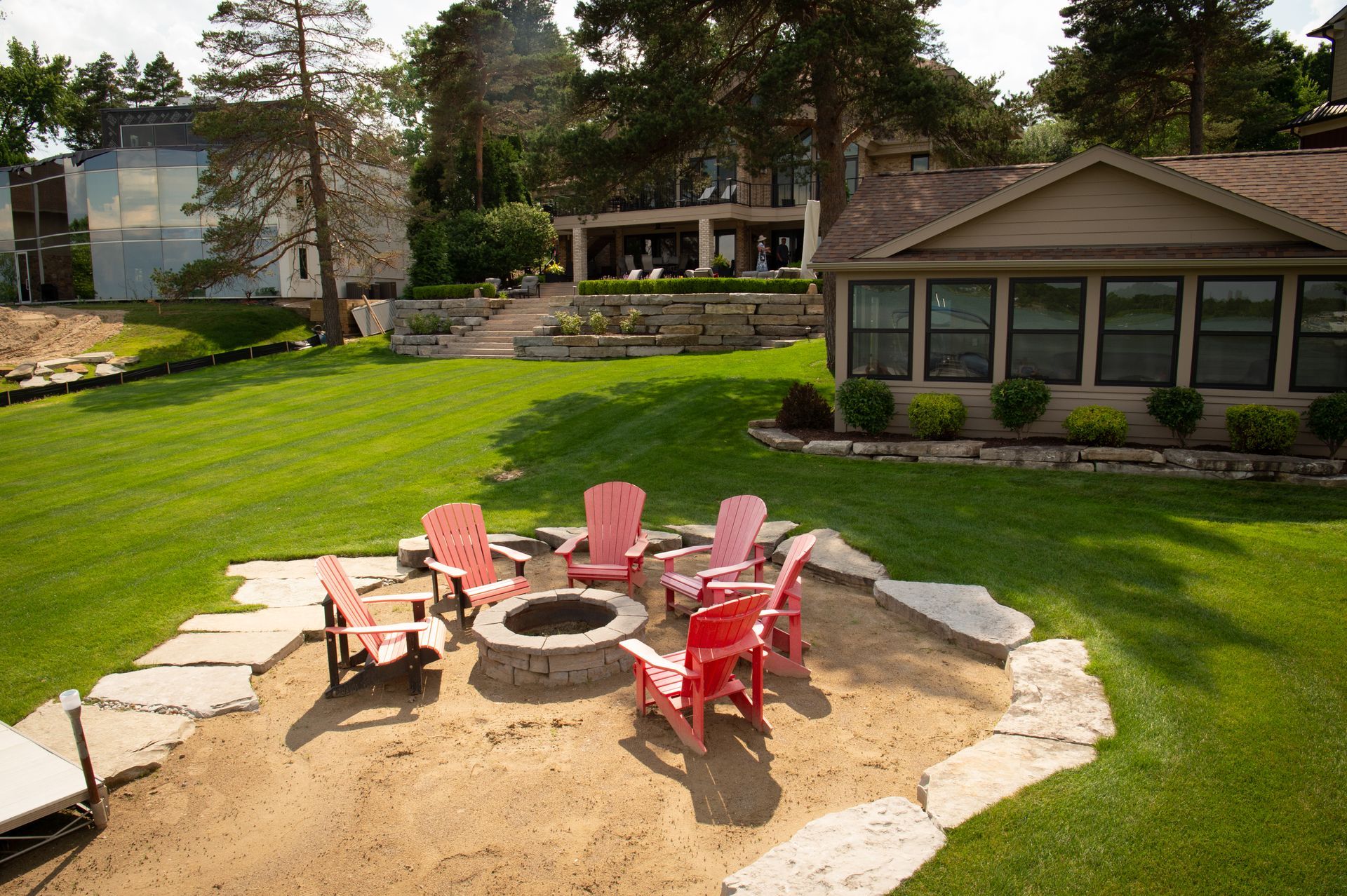 Fire pit with red chairs on a sandy area, surrounded by grass and a lakefront house.