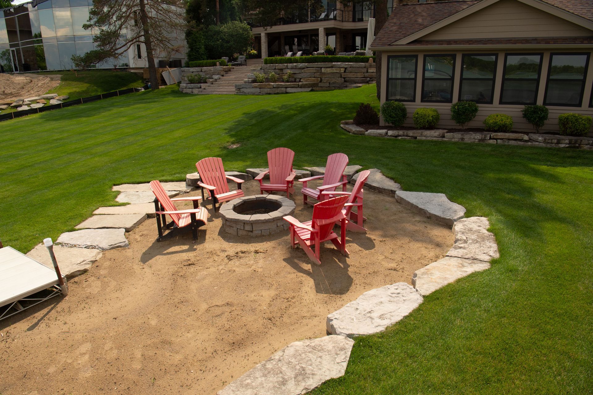 Fire pit with red and pink chairs on sand, surrounded by stone and green grass.