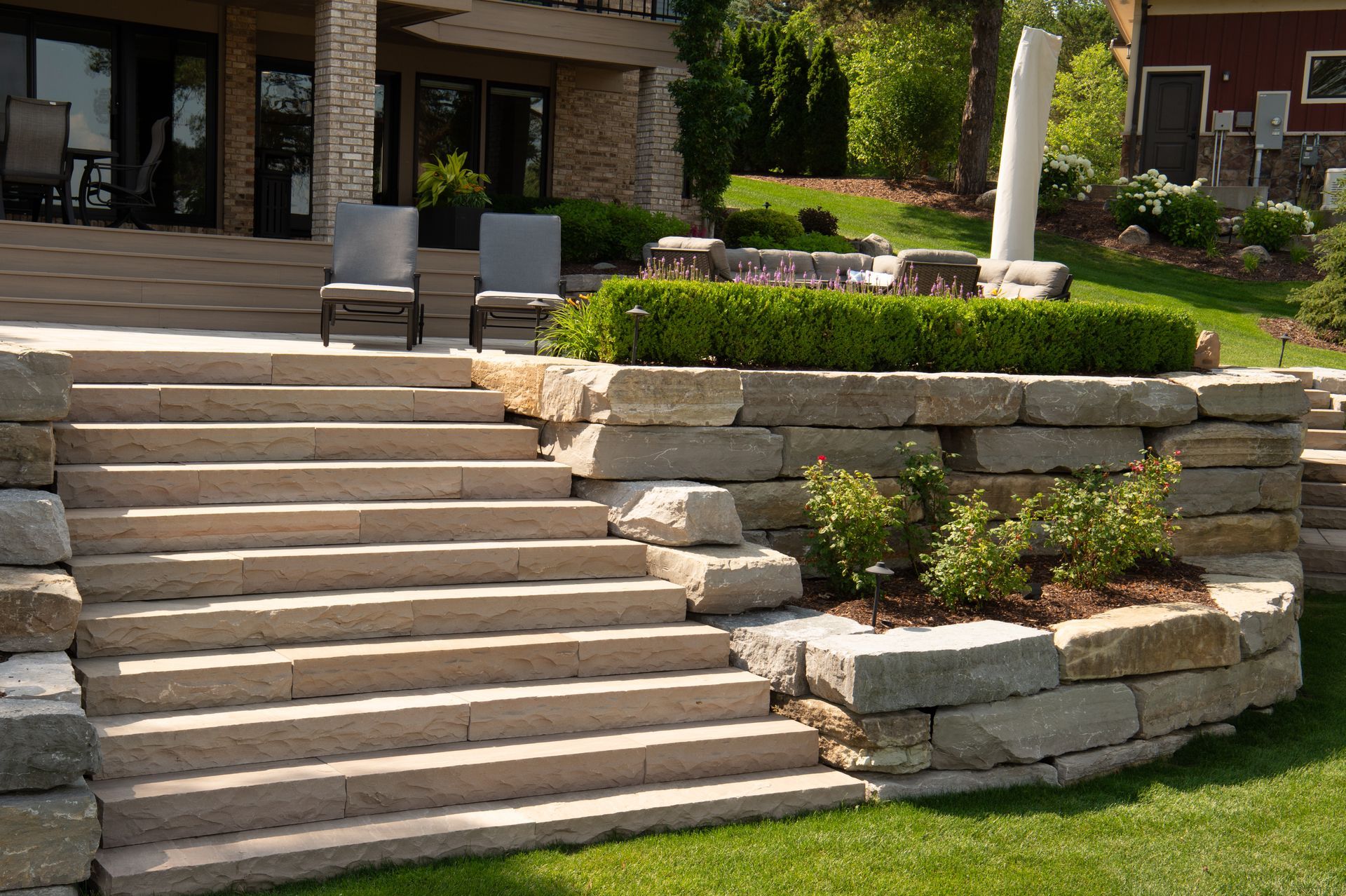Stone steps leading up to a patio with seating, surrounded by landscaping and lawn.