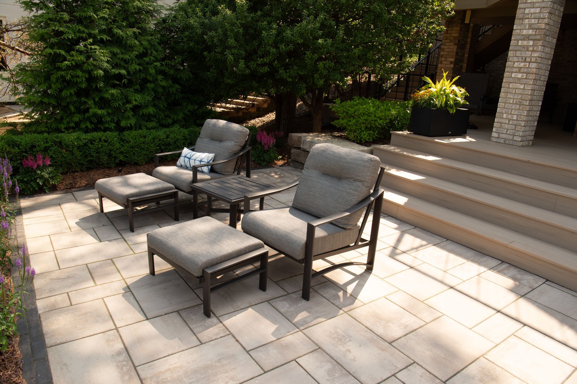 Two gray lounge chairs with ottomans on a paved patio, near greenery and steps.