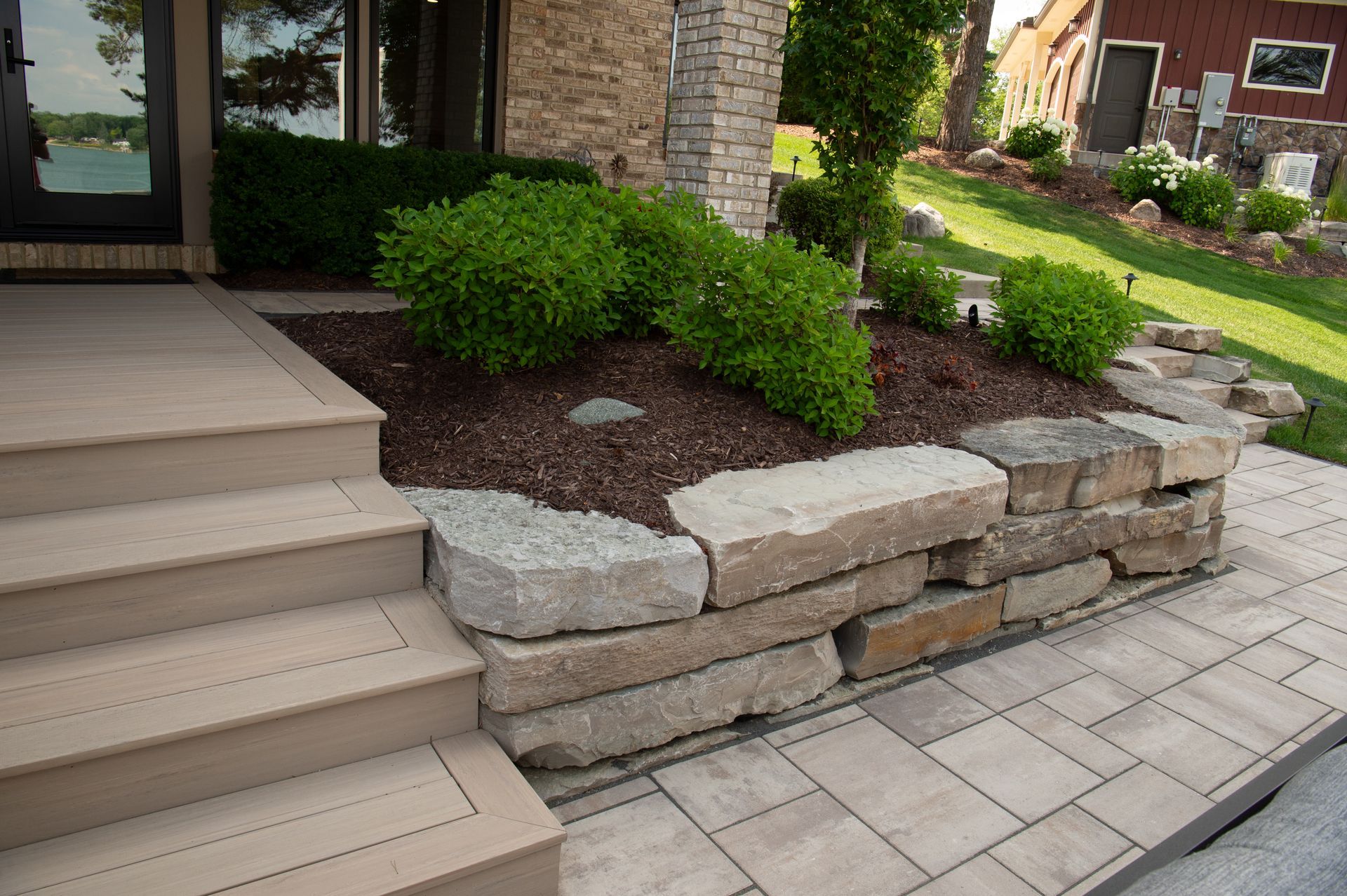Stone-walled planter bed with green shrubs and mulch near a building's entrance and light-colored steps.