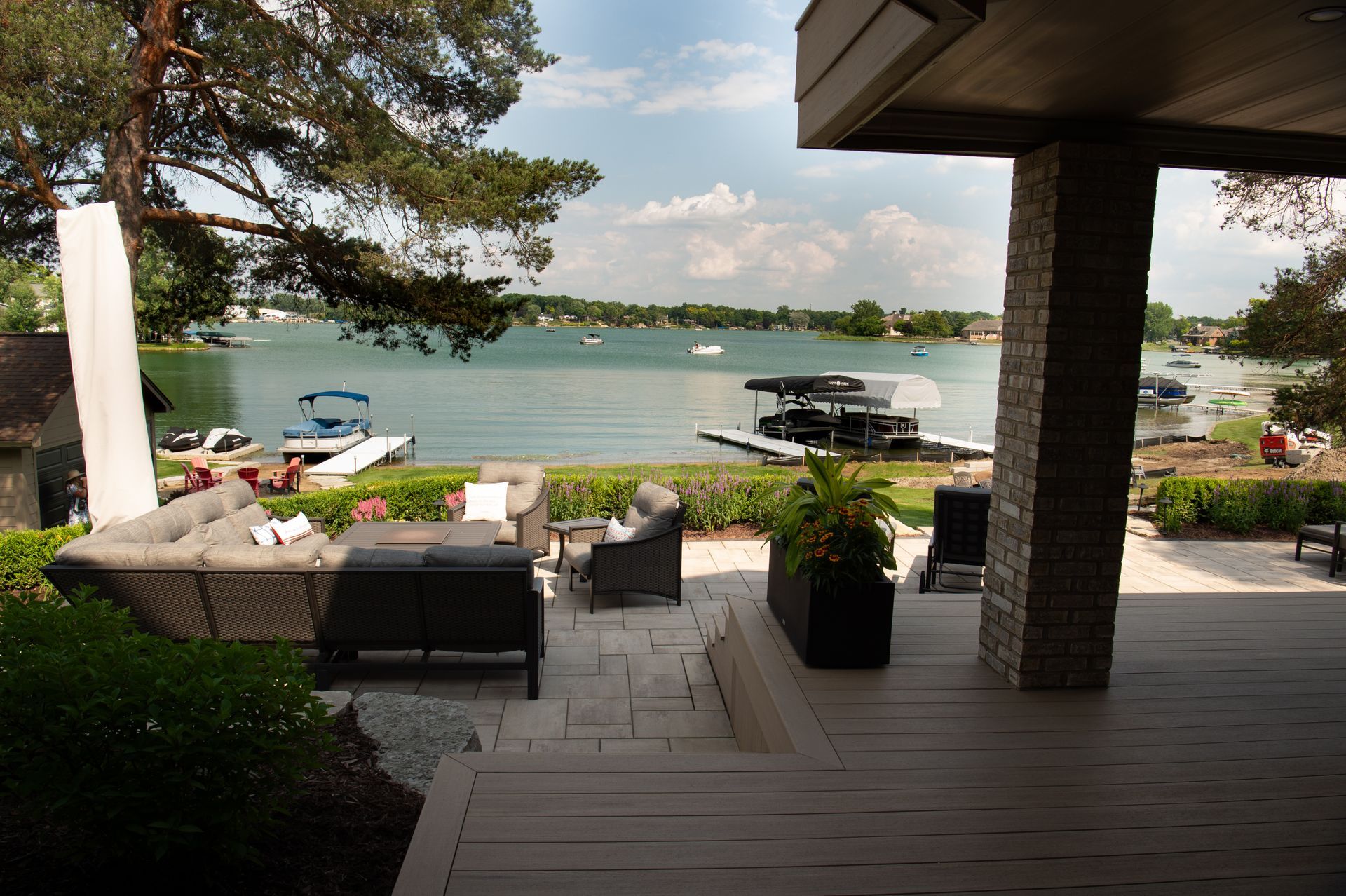 Outdoor patio overlooking lake, furnished with seating. Boats docked, trees and blue sky.