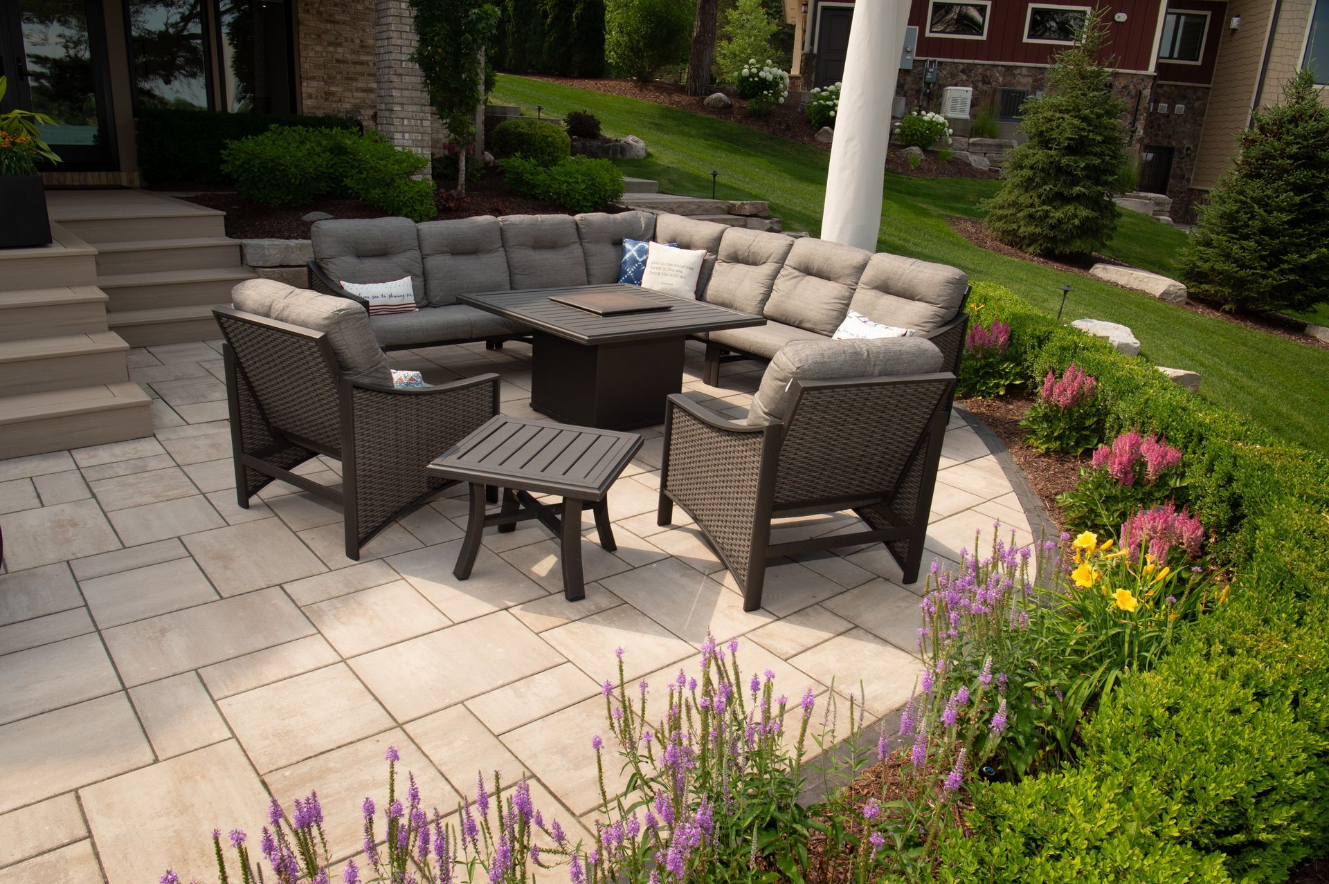 Outdoor seating arrangement on a patio: gray sectional, fire pit table, small side table, lush green landscaping.