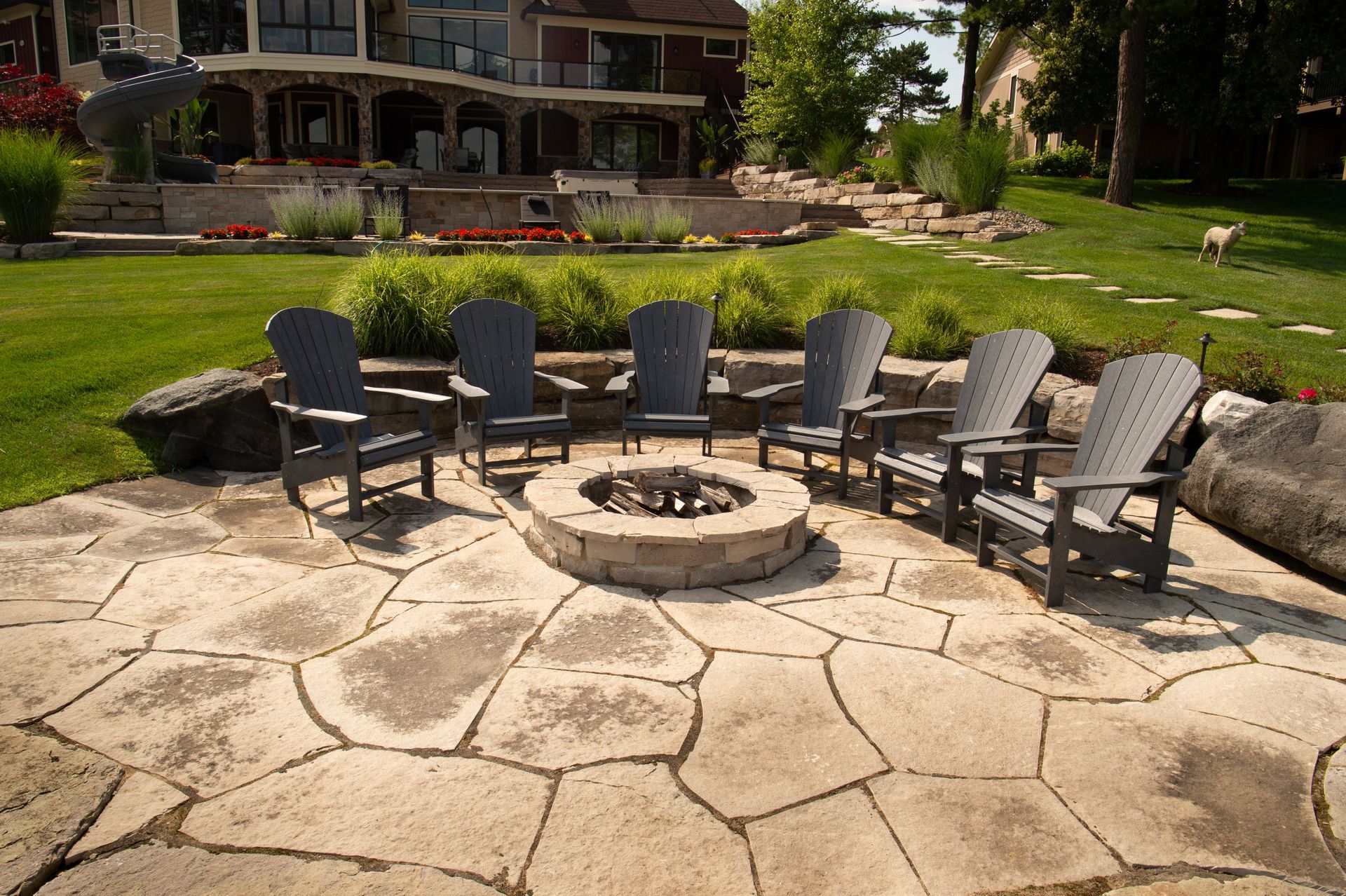 Outdoor seating area with fire pit and Adirondack chairs on a stone patio, with a grassy lawn.