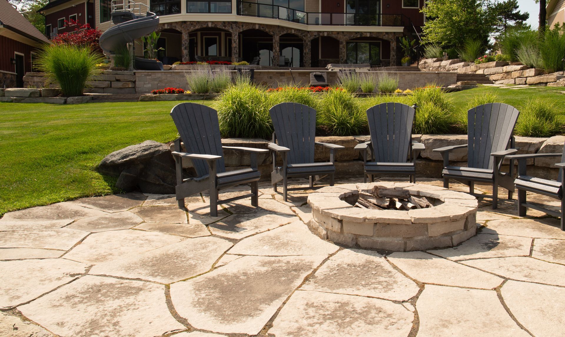 Stone patio with Adirondack chairs around a stone fire pit, overlooking a large house and green lawn.