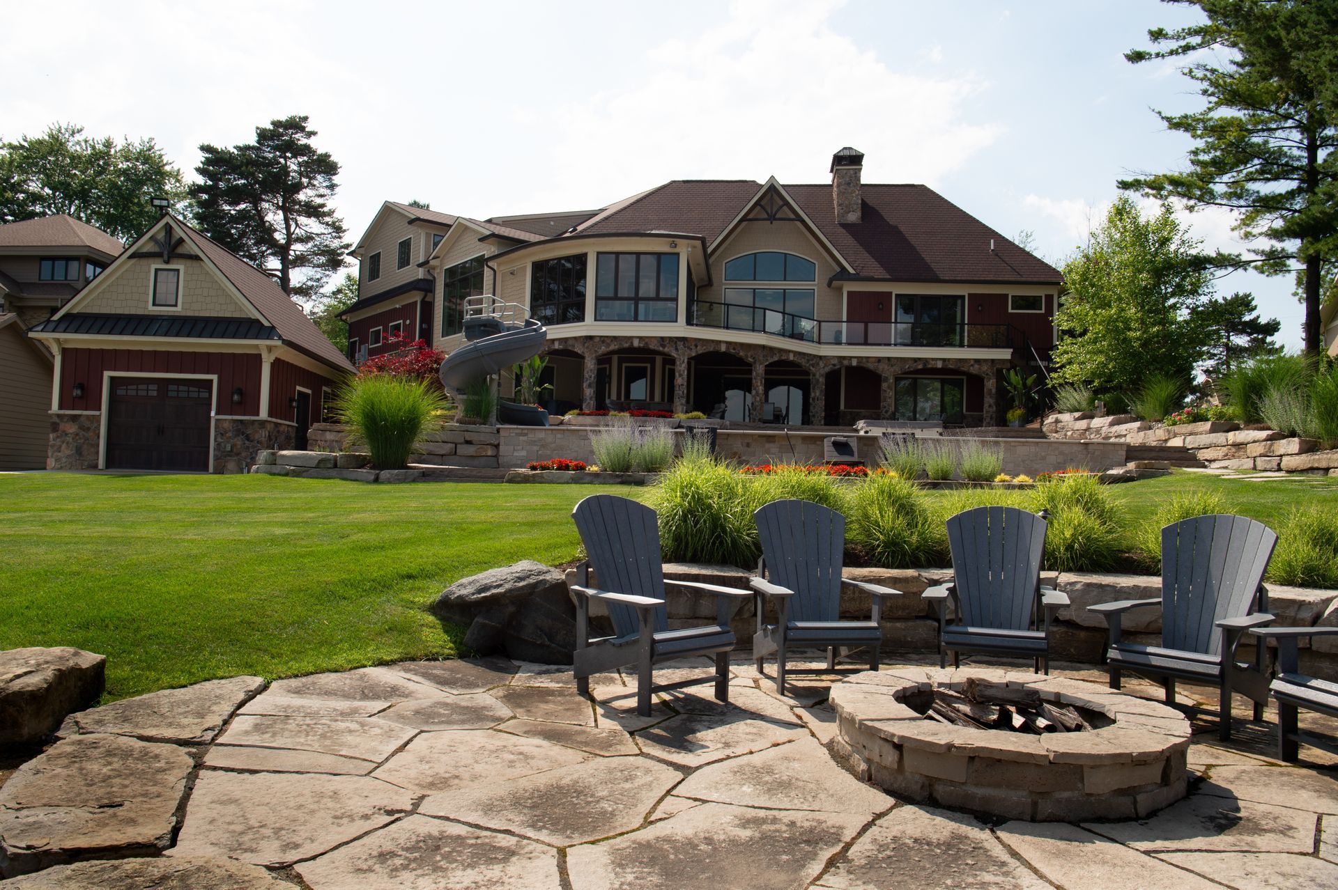 Large house with patio, fire pit, and Adirondack chairs on a green lawn. Sunny day.