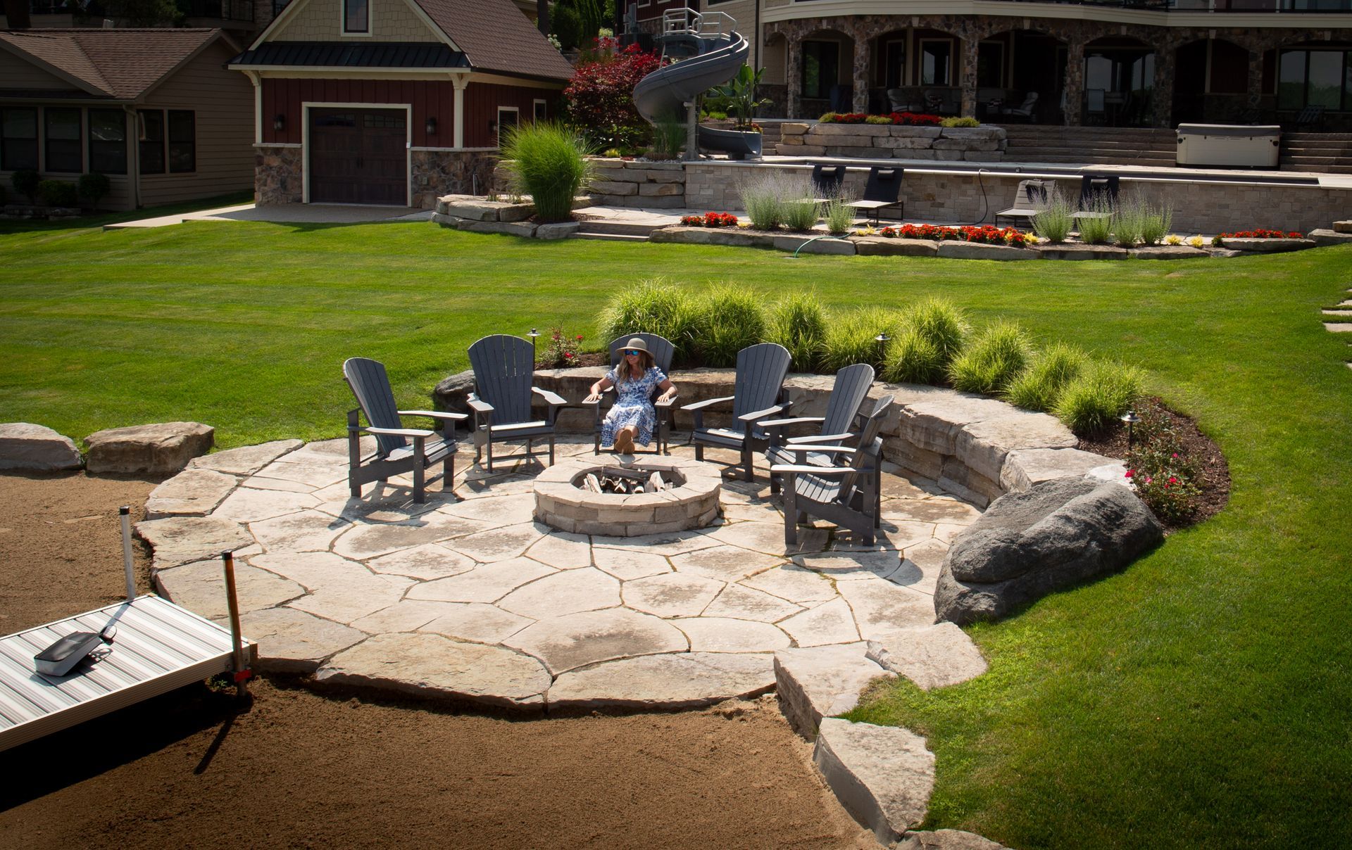 A patio with a fire pit surrounded by chairs, set in a grassy backyard near a lake.