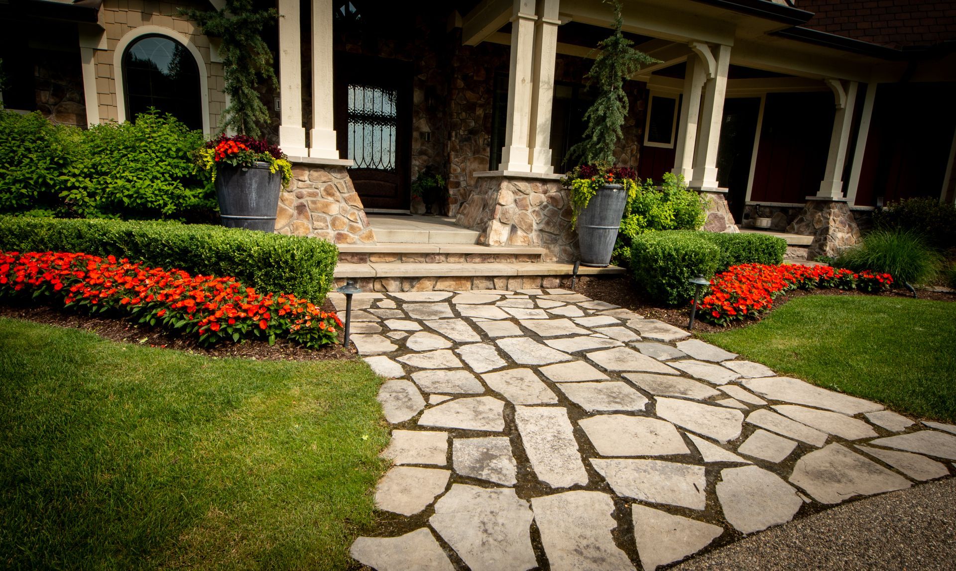 Stone walkway leading to a house with a porch. Landscaping includes red flowers, green shrubs, and grass.