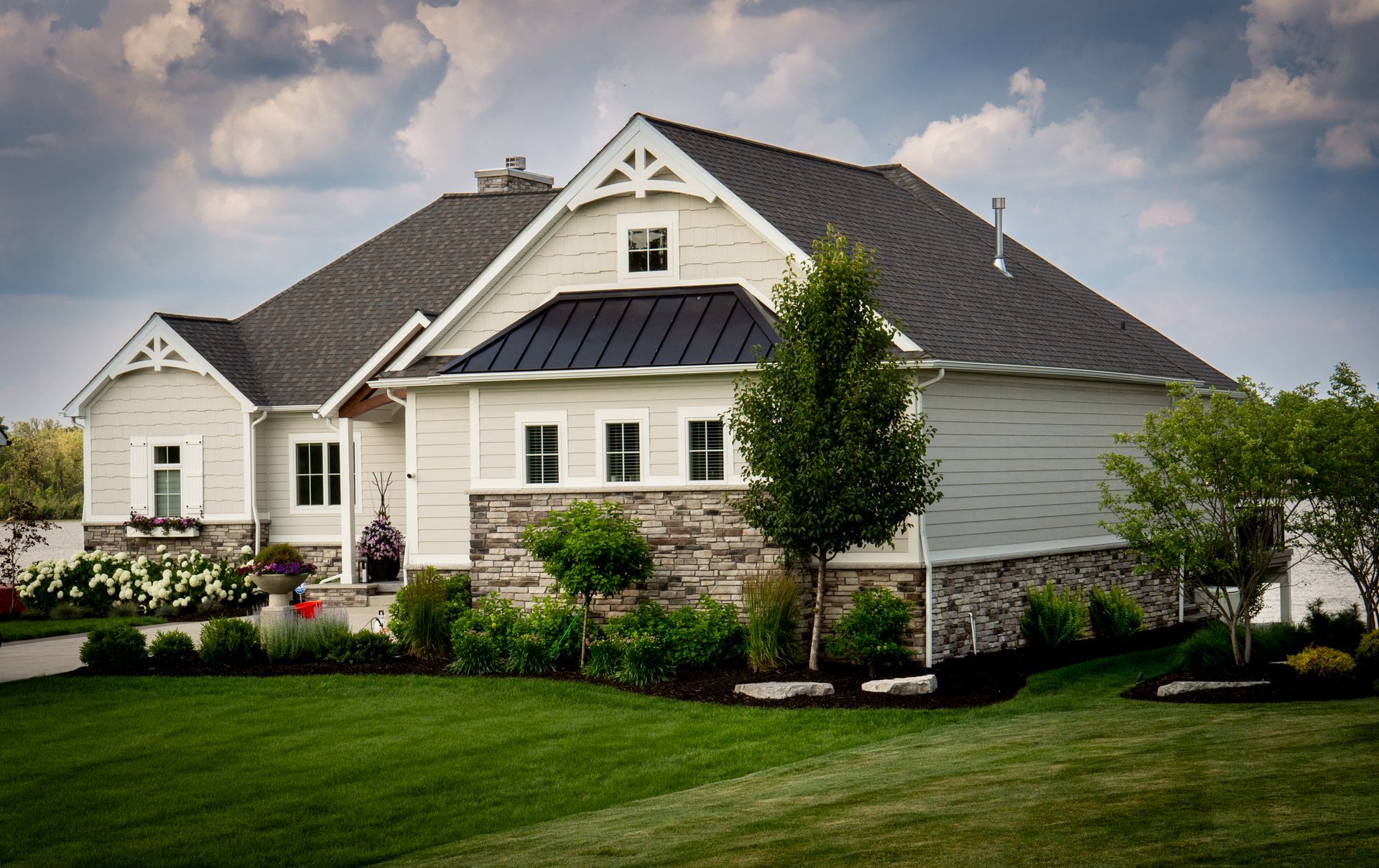 Beige house with gray roof, stone accents, and green lawn under a cloudy sky.