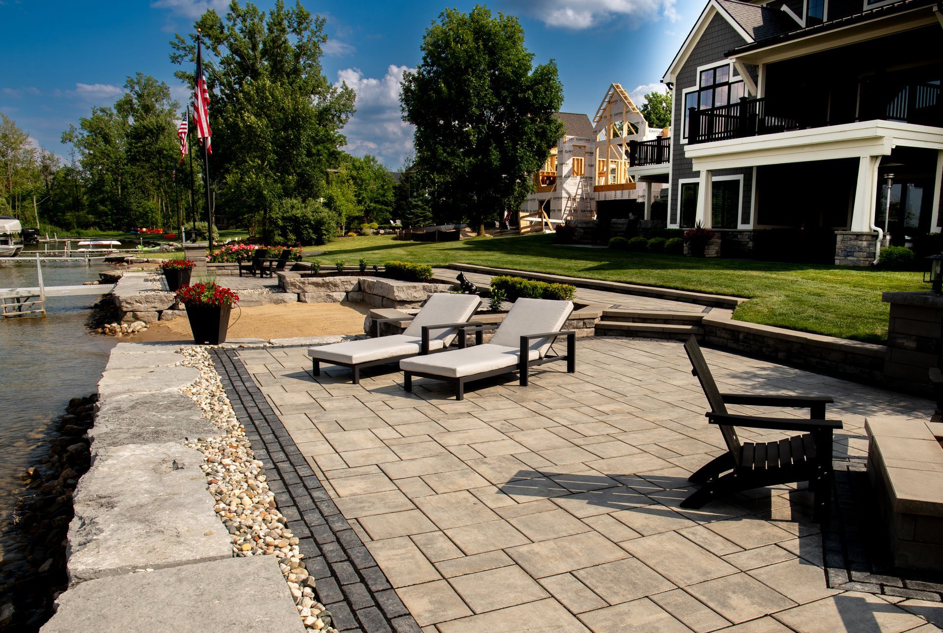 Lakeside patio with lounge chairs and lake view next to a large house, under a sunny sky.