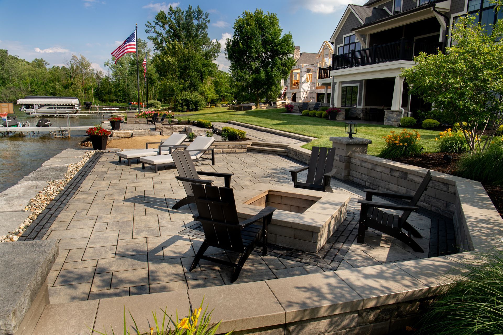 Stone patio with fire pit, lakefront, black Adirondack chairs, American flag, and large house.