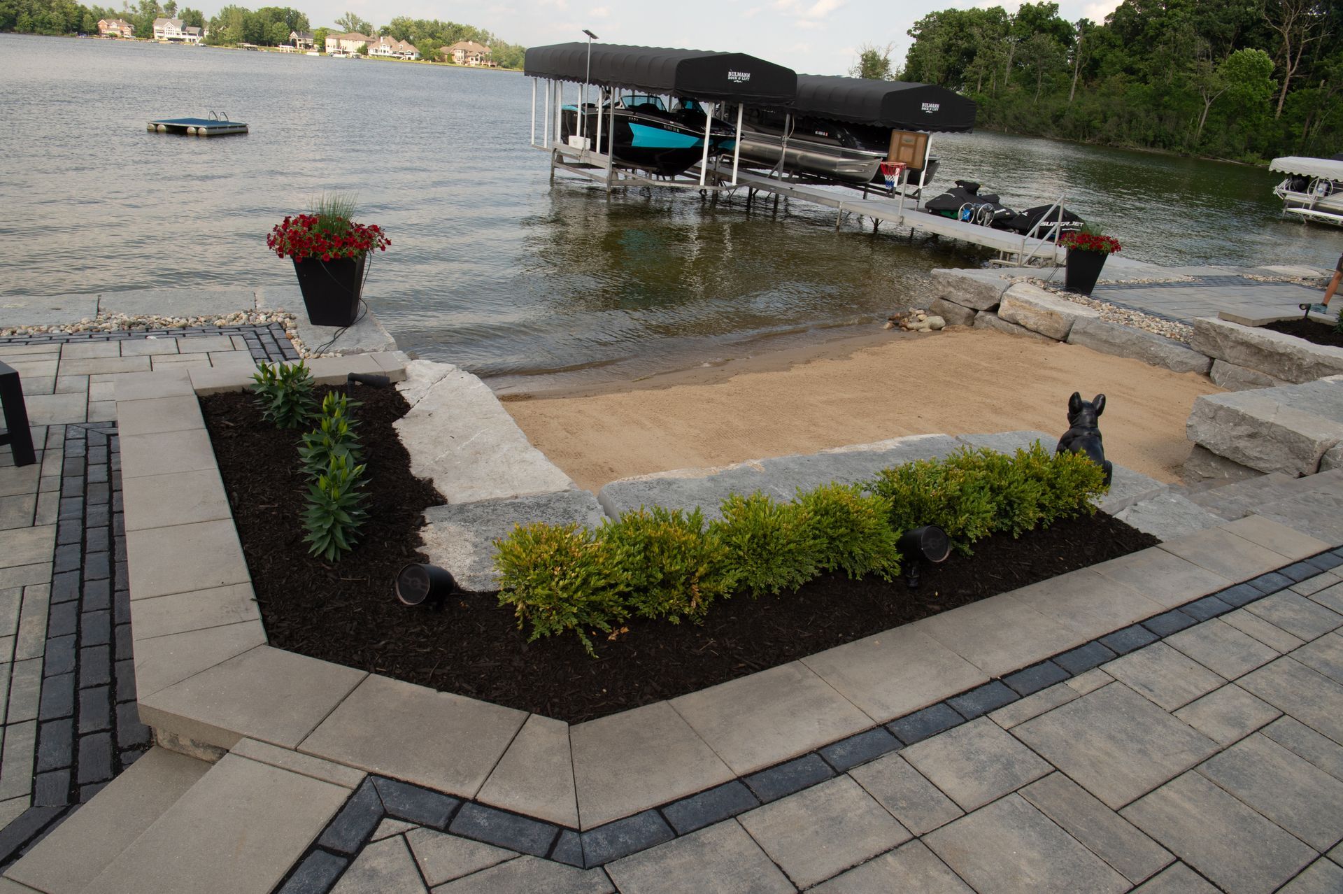 Patio with sandy area and lakefront dock. Pavers, landscaping, and a dark-hulled boat are visible.