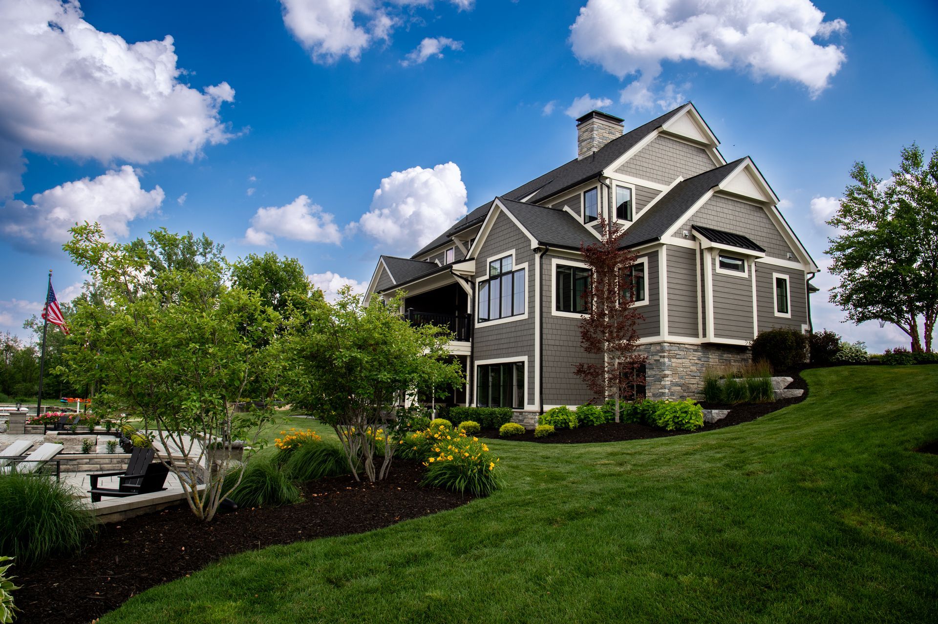 Gray and stone house on a grassy hill under a blue sky with fluffy clouds. Landscaping in foreground.