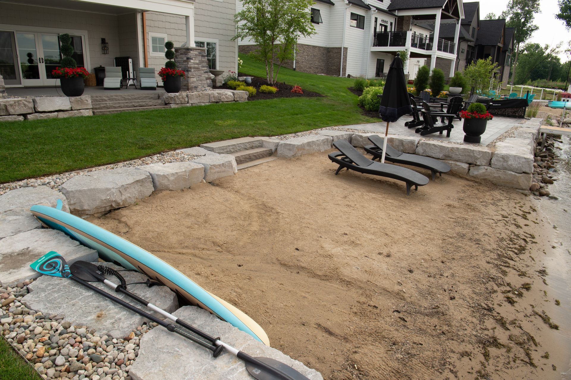 Sandy beach area with lounge chairs, kayak, and stone retaining walls near a lake.
