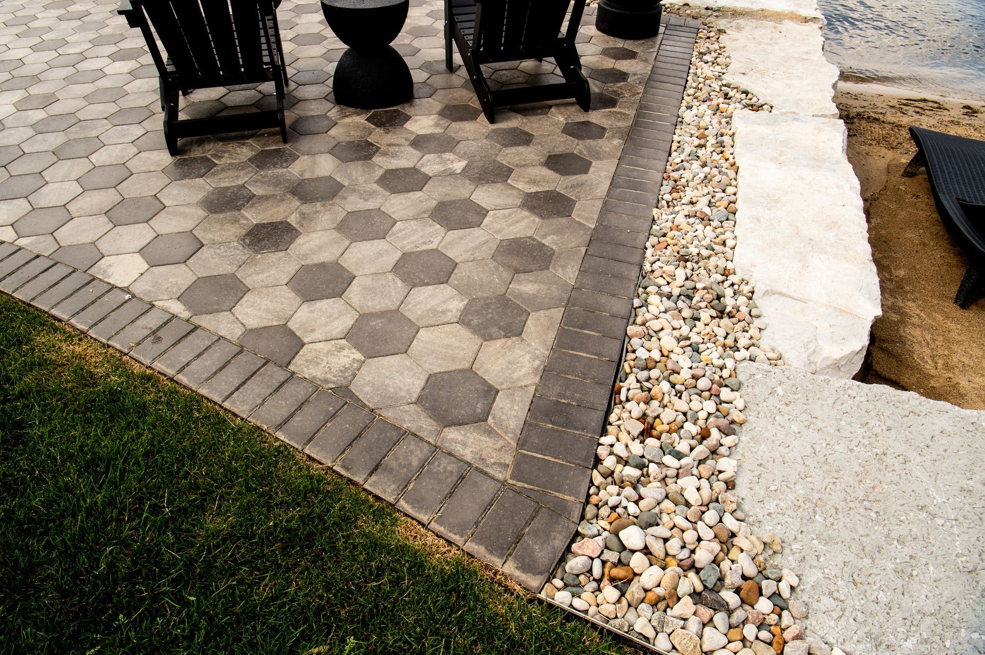 Patio with hexagonal pavers, stone border, gravel, and grass. Black chairs sit on the patio.