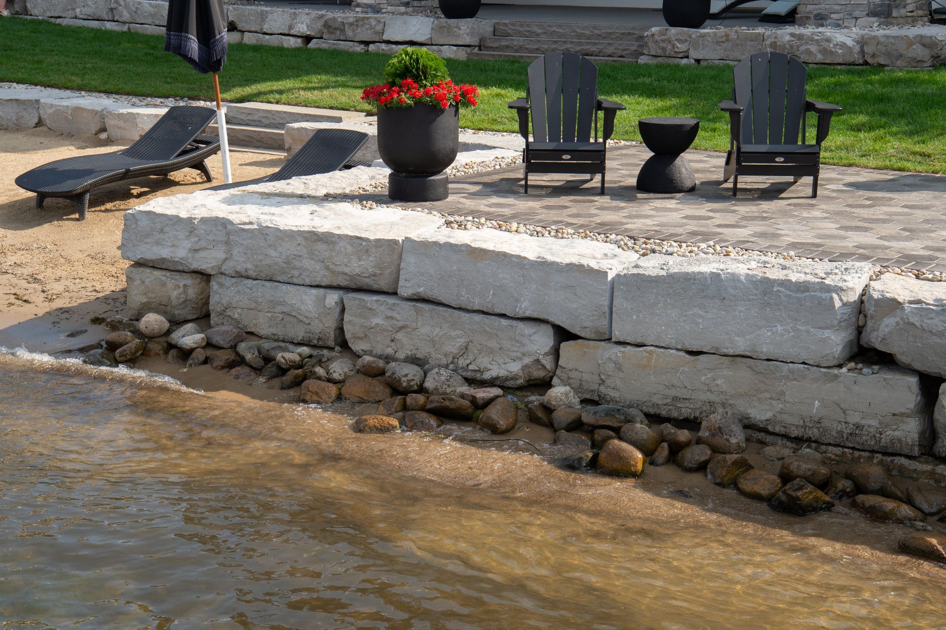 Stone retaining wall with a patio area and lake view. Two chairs, a table, and a flower pot are visible.