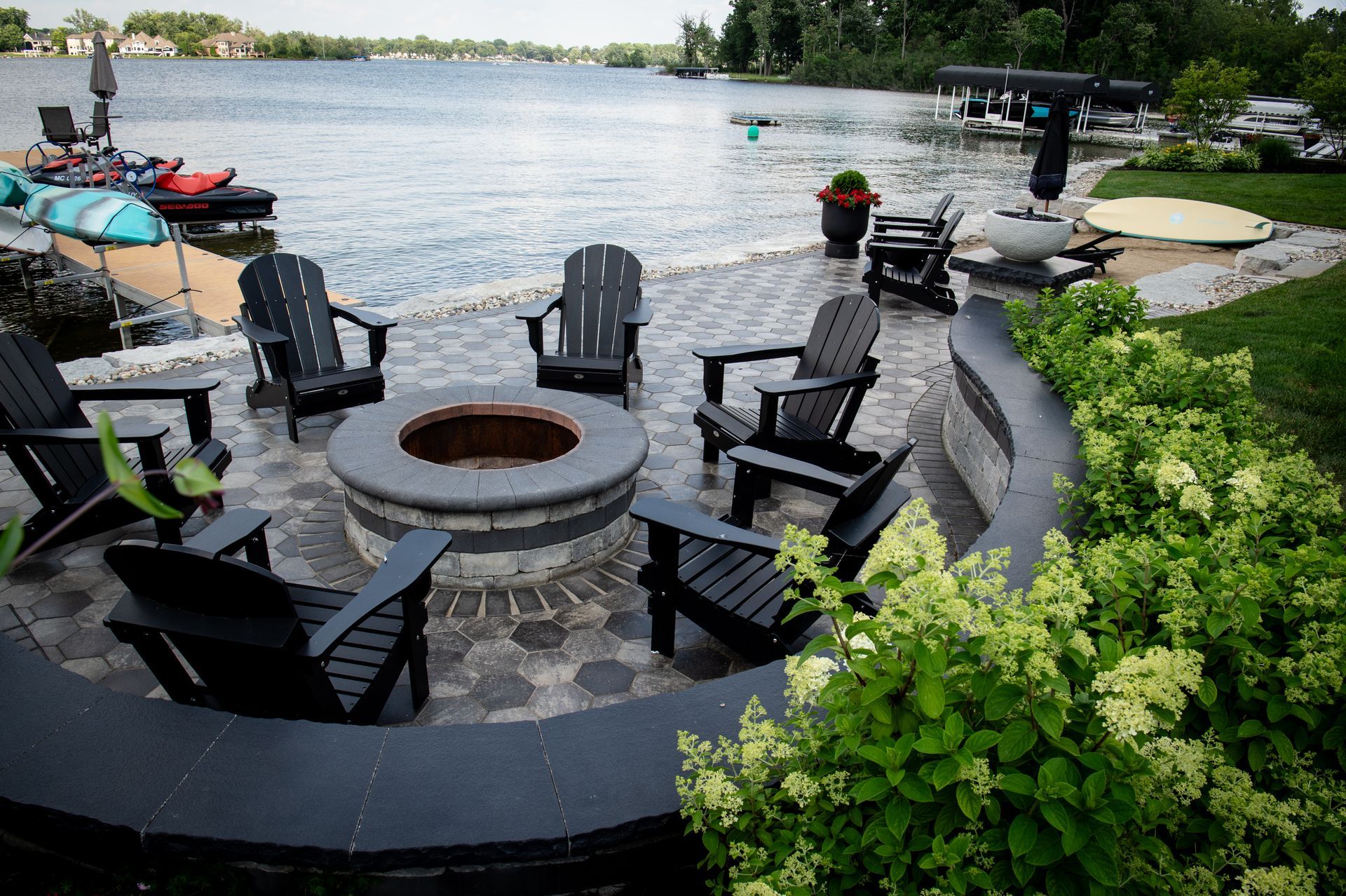 A lakeside patio with a fire pit surrounded by black Adirondack chairs.