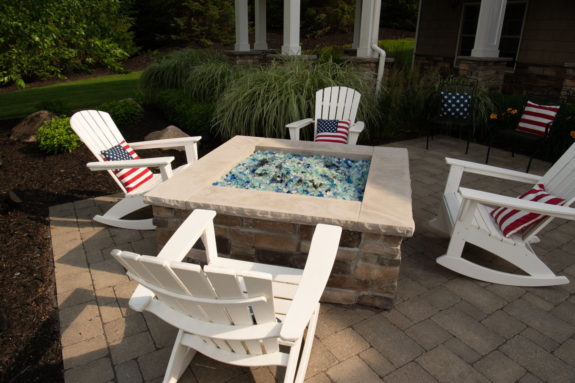 Four white rocking chairs around a stone fire pit on a patio, with American flag accents.