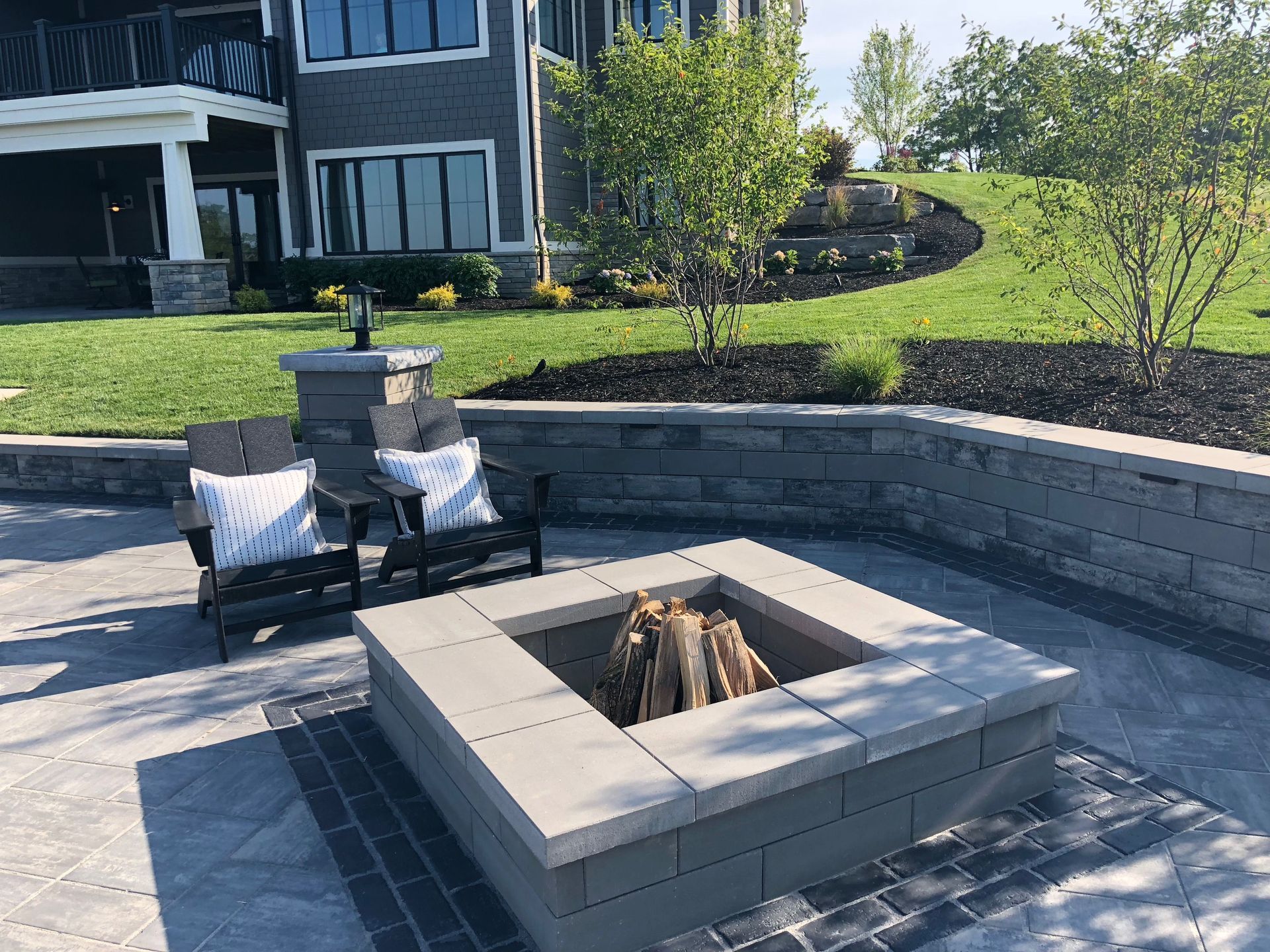 Outdoor patio with stone fire pit, chairs, and a retaining wall near a house.