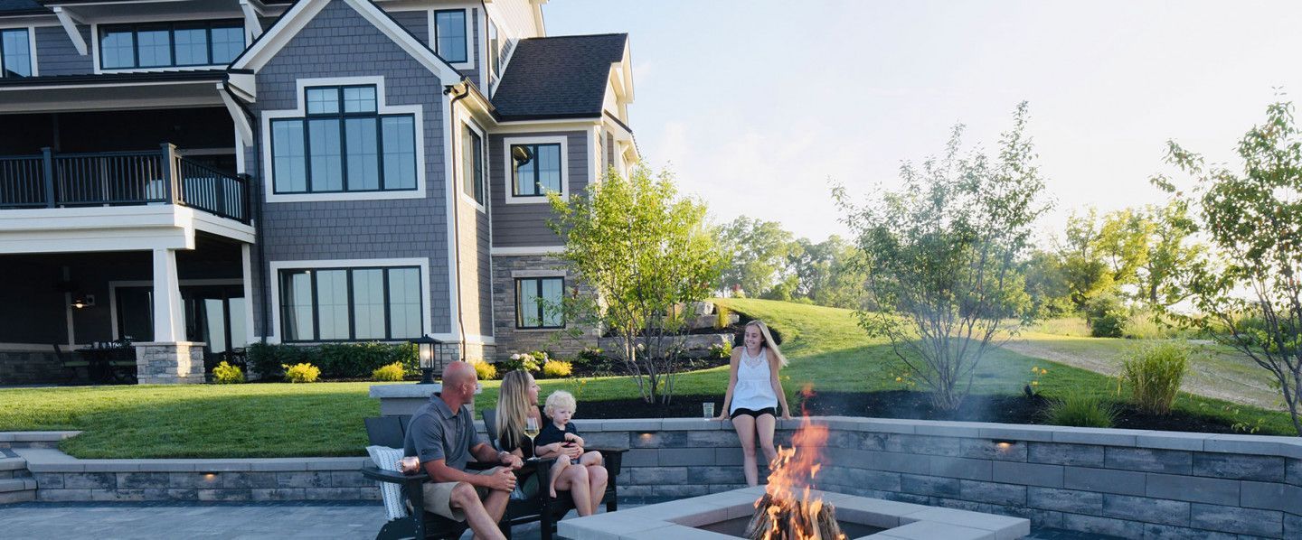 Family gathers around a fire pit in front of a large house.