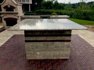 Outdoor granite-topped grilling island with brick and block base, on a brick patio, near an outdoor kitchen.