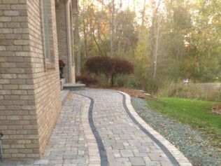 Brick pathway curves from a brick building's entrance, bordered by grass and small rocks, with trees in the background.