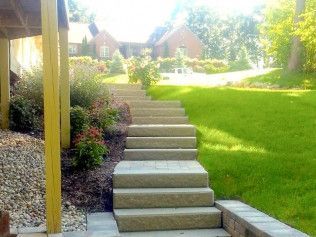 Stone steps lead up a grassy hill to a brick house. Green trees and plants surround the steps.