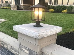 Stone pillar with a lit lantern in front of a house, set in grass and landscaping.