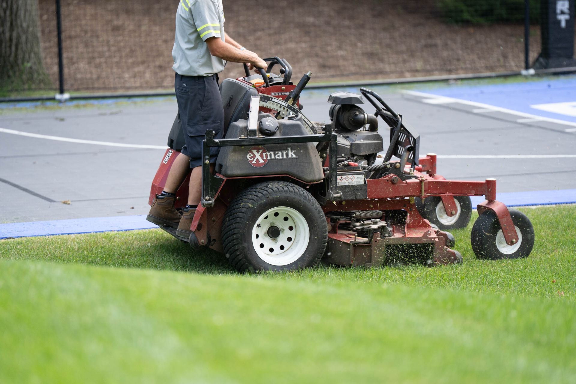 A person mowing a green lawn with a red Exmark riding mower next to a blue and white court.